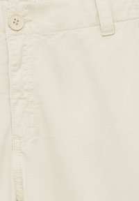 Gros plan sur un tissu beige clair, présentant une texture douce, des coutures visibles et une fermeture à bouton unique sur un pantalon.