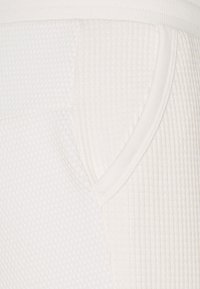 Witte gestructureerde stof met een ribdesign en een zijzak. Vlotte afwerking rond de tailleband. Focus op gedetailleerde stitching.