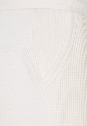 Witte gestructureerde stof met een ribdesign en een zijzak. Vlotte afwerking rond de tailleband. Focus op gedetailleerde stitching.