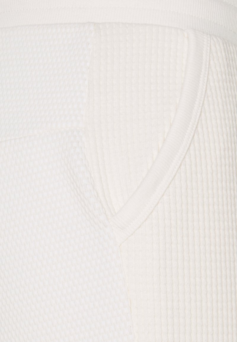 Witte gestructureerde stof met een ribdesign en een zijzak. Vlotte afwerking rond de tailleband. Focus op gedetailleerde stitching.