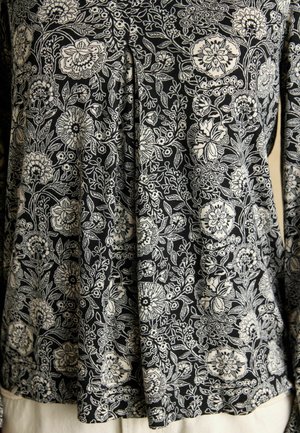 Čierna tkanina s hustým bielym kvetinovým vzorom, ktorý zahŕňa rôzne kvety, listy a vinič, nosená ako top s dlhým rukávom.