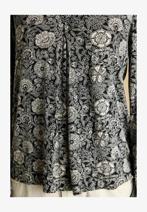 Čierna tkanina s hustým bielym kvetinovým vzorom, ktorý zahŕňa rôzne kvety, listy a vinič, nosená ako top s dlhým rukávom.