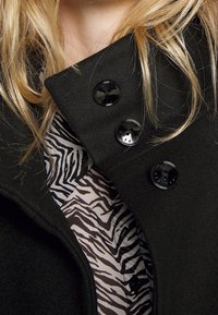 Cappotto di lana nero con colletto alto, dotato di tre bottoni neri. Il tessuto interno presenta un motivo a strisce zebra in marrone e crema.