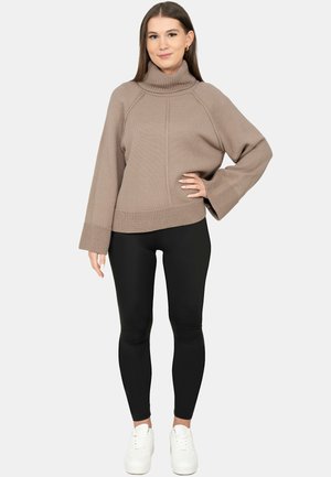 Woman standing wearing a beige turtleneck sweater, black leggings, and white sneakers, with one hand on her hip.