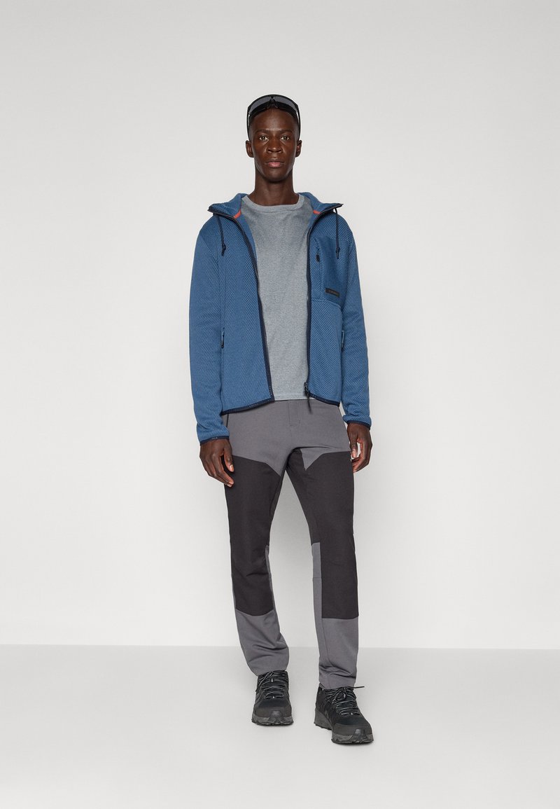 Blau strukturierte Zip-Jacke, graues Rundhals-T-Shirt, graue und schwarze Hose mit Panelelementen, schwarze Wanderschuhe, insgesamt Outdoor-Bekleidung.