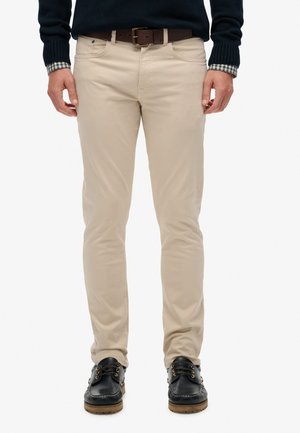 Hombre con pantalones beige ajustados, cinturón oscuro, suéter azul marino sobre una camisa de cuadros y zapatos negros con cordones, de pie contra un fondo blanco.