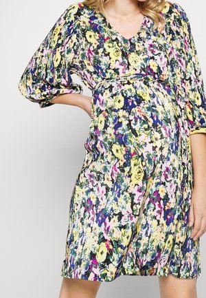 Mujer con vestido hasta la rodilla con estampado floral con flores amarillas, rosas y moradas y mangas abullonadas hasta tres cuartos, mano apoyada en la cadera.
