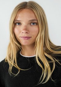 Zwarte gebreide trui met een ronde hals, gedragen over een wit shirt. Lange, rechte blonde haren omlijsten het gezicht. Lichte huidskleur.