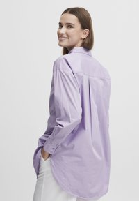 Light purple button-up shirt with long sleeves, rounded hem, and a back pleat. Fabric appears smooth and lightweight. Worn with white pants.