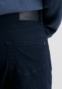 Nahaufnahme der dunkelblauen Jeans mit einer hinteren Tasche, auf der die sichtbaren Nähte und ein schwarzes Lederetikett mit dem Markennamen "Pierre Cardin" zu sehen sind.
