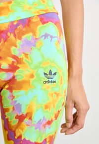 Farebné legíny s technikou tie-dye v purpurovej, zelenej, oranžovej a žltej látke, s čiernym logom Adidas na ľavom stehne.