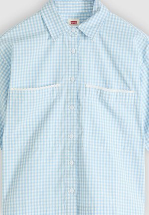 Chemise à manches courtes en vichy bleu clair et blanc avec des boutons blancs, un col pointu, deux poches avant bordées de dentelle blanche.