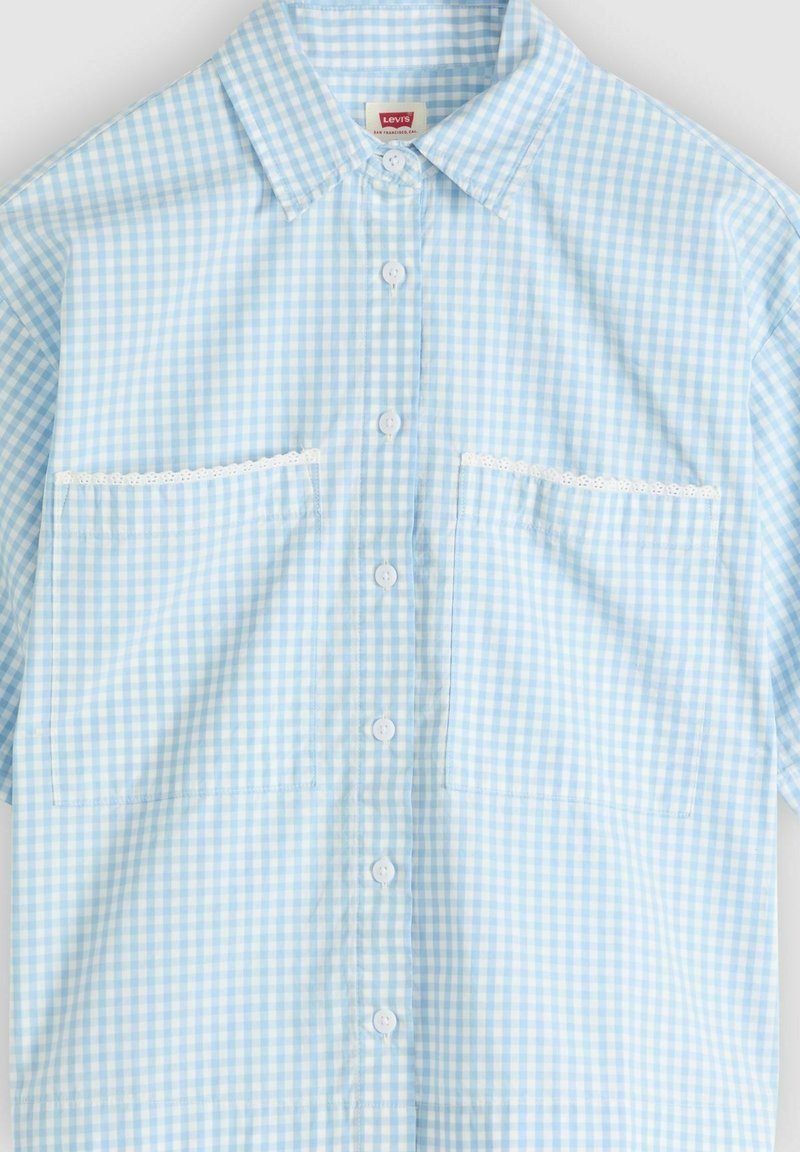 Chemise à manches courtes en vichy bleu clair et blanc avec des boutons blancs, un col pointu, deux poches avant bordées de dentelle blanche.