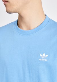 Camiseta de algodón de color azul claro con escote redondo y pequeño logotipo bordado de Adidas en blanco en el lado izquierdo del pecho. Textura suave.