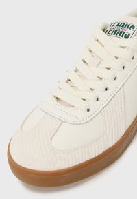 Beli čevelj z belim snežkom, ki ima teksturiran korduroj in gladko usnjeno zgornjo plast, okrogle vezalke ter gumijast podplat. Lacoste logotip na jeziku.