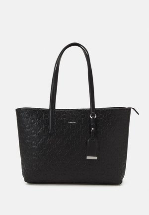 Sort Calvin Klein tote taske med præget CK-logo mønster, to lange håndtag og vedhæftet rektangulært mærke med sølvdetalje.