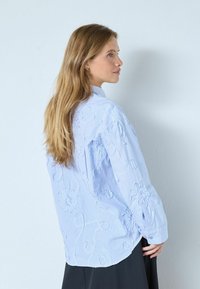 Vaaleansininen, raidallinen puuvillapaita, jossa on kukkaistikkaus, pitkät hihat napillisine kalvosimineen ja klassinen kaulus. Sileä kangas ja rento istuvuus.