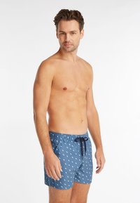 Shorts de bain bleus avec un motif de feuilles blanches, taille élastique et cordon de serrage. Le tissu semble léger et lisse. Le modèle se tient de face.