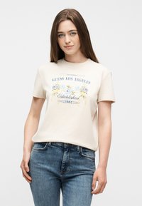 Νεαρή γυναίκα με μακριά καστανά μαλλιά που φοράει κοντομάνικη μπλούζα με κρεμ χρώμα και λουλουδένιο σχέδιο με την επιγραφή «Guess Los Angeles Established 1981» και μπλε τζιν.