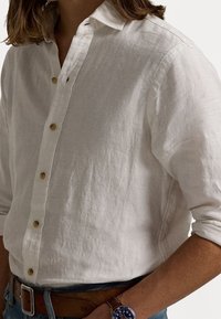 Lněná košile na knoflíky v krémové barvě s jemnou texturou, s límečkem do špičky a hnědými knoflíky. Kombinováno s modrými džíny a hodinkami.