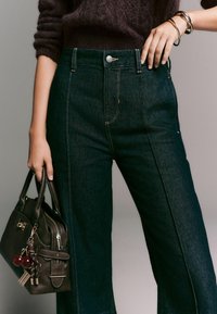 Dunkelblaue, hochtaillierte Jeans mit kontrastierenden Nähten und einem Vorderknopfverschluss. In der Hand wird eine kleine braune Handtasche mit Anhängern gehalten.