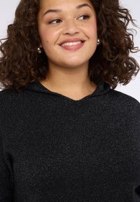 Felpa nera con una sottile texture scintillante, dotata di cappuccio e vestibilità comoda. Primo piano della scollatura e della parte superiore del capo.