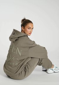 Sudadera oliva de gran tamaño con detalles de logotipo texturizado, combinada con joggers a juego. Presenta un corte relajado y material de algodón suave.