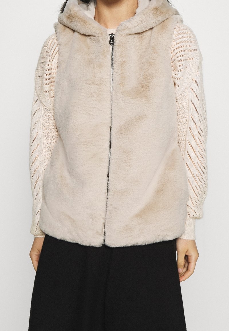 Faux fur-väst i ljusbeige, med huva, dragkedja och texturerad yta. Bärs över en vit stickad långärmad topp.