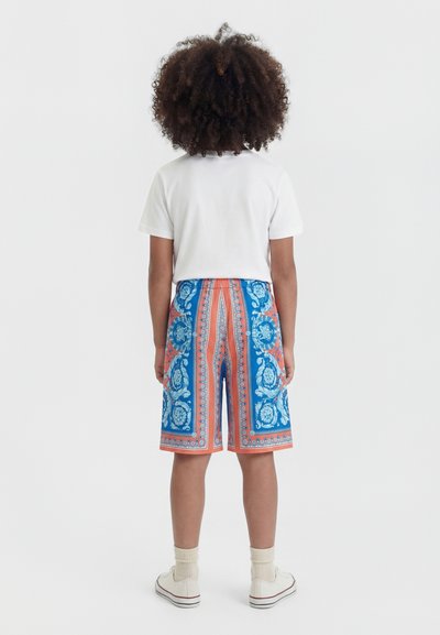 Παιδί με μαλλιά σε μπούκλες στέκεται πλάτη, φορώντας λευκό t-shirt, πολύχρωμο σορτσάκι με μοτίβα μέχρι το γόνατο, λευκές κάλτσες και λευκά αθλητικά παπούτσια.