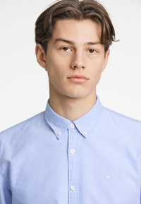 Camicia azzurra chiara con colletto ripiegato, dotata di bottoni bianchi e di un piccolo emblema ricamato sul lato sinistro. Tessuto dalla consistenza liscia.