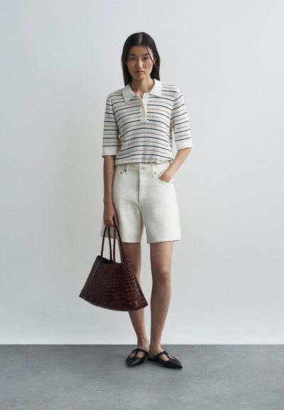 Īsām piedurknēm, baltām svītrām adīts polo krekls ar apkakli, pārī ar baltām džinsu šortiem. Tur rokās tumši sarkanu austu somu un valkājot melnas čības.