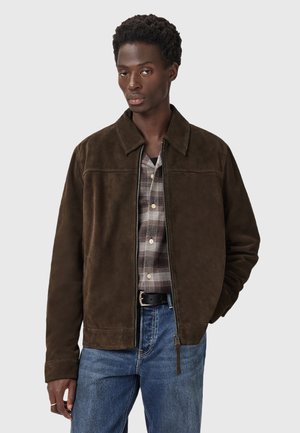 Hombre con una chaqueta de ante marrón oscuro sobre una camisa de cuadros, jeans azules y un cinturón negro, de pie frente a un fondo claro y liso.