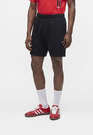 Muž v čiernych krátkych nohaviciach, bielych členkových ponožkách a červených teniskách Adidas s bielymi pruhmi, stojí na jednoliatom bielom pozadí.