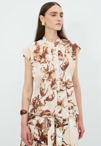 Blusa senza maniche con stampa floreale in tonalità crema e marrone, caratterizzata da undavanti plissè, chiusura con bottoni e vestibilità comoda. Tessuto dalla texture liscia.