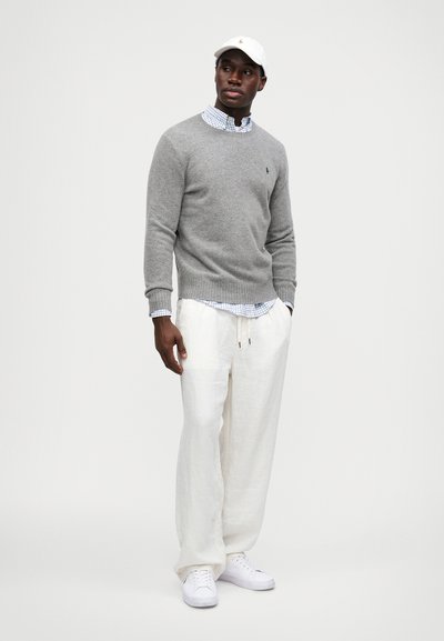 Pullover de lã cinza sobre uma camisa xadrez azul, combinado com calças de linho brancas e ténis brancos, complementado por um boné branco.