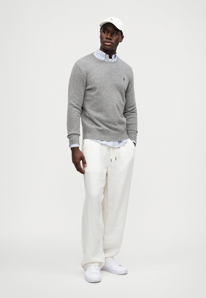 Suéter de lana gris sobre una camisa a cuadros azul, combinado con pantalones de lino blancos y zapatillas blancas, complementado con una gorra blanca.