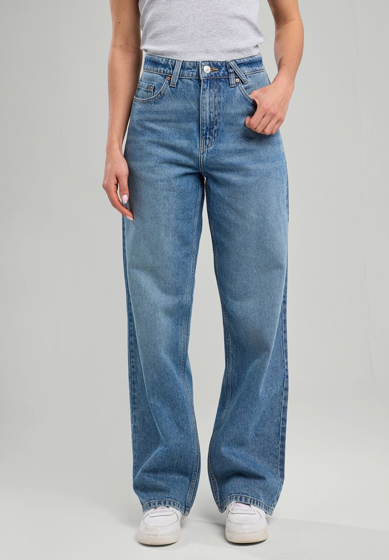 Jeans de tiro alto en azul claro con un ajuste holgado, que presentan cinco bolsillos, passadores para cinturón y un desvanecimiento sutil en la tela. Combinados con zapatillas blancas.