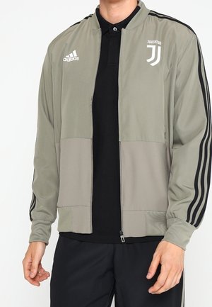 Henkilö, joka käyttää beigeä Adidas-urheilutakkia, jossa on mustat raidat ja Juventuksen logo, mustan poolopaidan ja mustien housujen päällä.