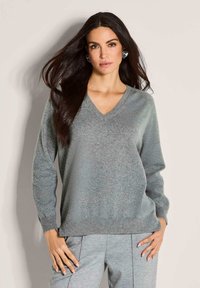 Frau mit langen, dunklen Haaren trägt einen grauen V-Ausschnitt-Pullover und passende Hose, steht vor einem schlichten beigen Hintergrund, mit Händen in die Hüften gestützt.