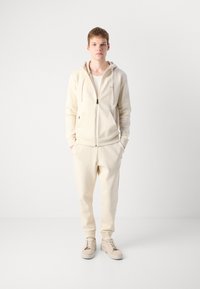 Ung mand står med hænderne i lommerne iført beige hoodie, matchende joggers, hvid skjorte og beige sneakers mod en hvid baggrund.
