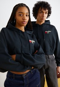 Svart bomullshoodies med en forkortet design. Den ene har et rødt rosegrafikk og logo. Begge modellene har på seg høyhøydejeans i kontrastfarger.
