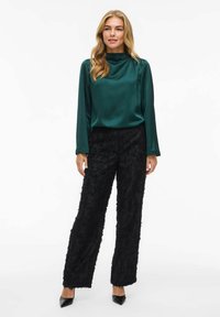 Grüne Satin-Bluse mit langen Ärmeln und hohem Kragen, kombiniert mit schwarzen, strukturierten weit geschnittenen Hosen mit einem flauschigen Muster und spitzen schwarzen Absätzen.