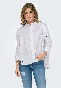 Camicia leggera bianca con ricami a forma di cuore rossi e blu. Presenta un colletto, chiusura frontale con bottoni e maniche lunghe dalla vestibilità ampia.