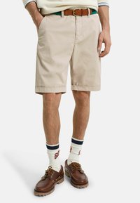 Beige katoenen shorts met zijzakken, omgeslagen zoom en een bruine leren riem. Bruine veterschoenen en witte sokken met blauwe strepen maken de look af.