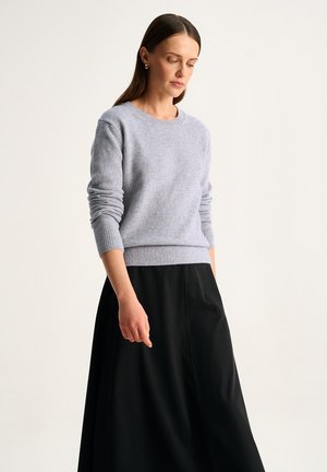 Kobieta o prostych brązowych włosach, ubrana w szary sweter z długim rękawem i czarną spódnicę, patrzy w dół z zrelaksowaną postawą na jednolitym tle.