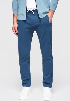 Uomo con pantaloni jogger blu con coulisse bianca, felpa con cappuccio azzurro con cerniera, e scarpe da ginnastica bianche, in piedi con una mano in tasca.