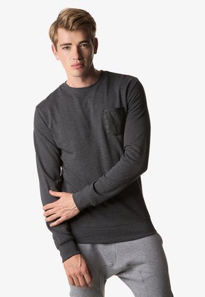 Grijze langärmige sweatshirt met een zwarte leren zak, ribbelmanchetten en -zoom, gemaakt van zachte stof. Model poseert met de armen gekruist.