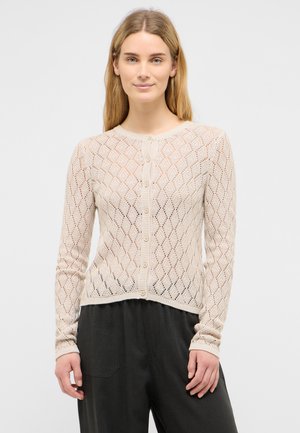 Woman with long blonde hair wearing a beige patterned button-up cardigan and black pants, standing against a plain light background.