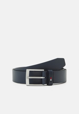 Ceinture en cuir noir avec une boucle rectangulaire argentée et un petit logo rouge, blanc et noir sur la boucle de la ceinture.