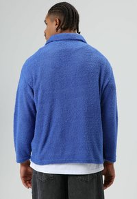 Blauwe fleece trui met een kraag, met een gestructureerd, pluizig oppervlak. Het ontwerp heeft lange mouwen en een losse pasvorm, gecombineerd met donkere shorts.